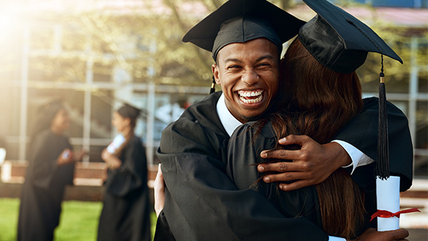 estudiante afroamericano sonriente con toga de graduación abrazando a una compañera