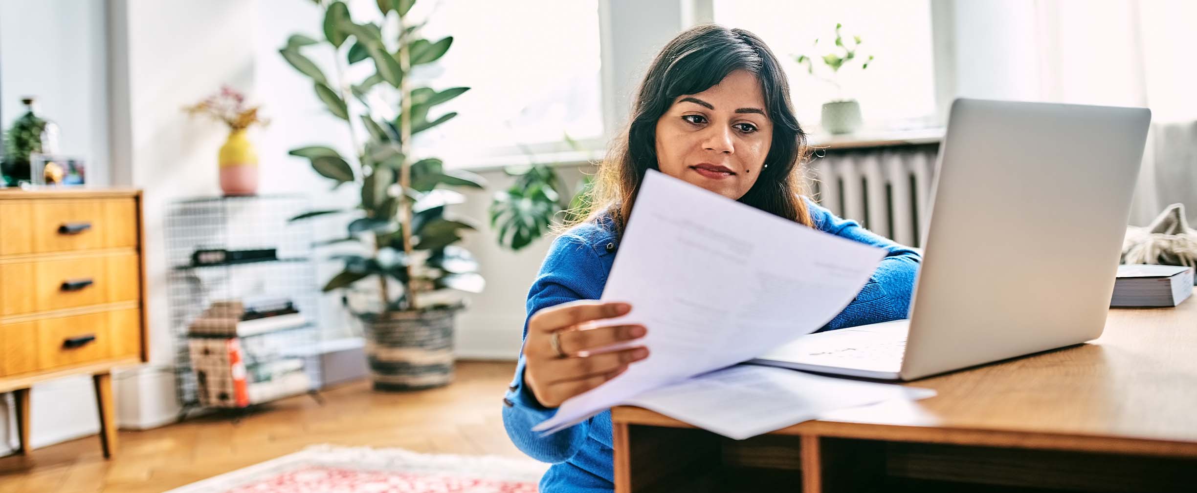 Una mujer revisa las finanzas utilizando una computadora portátil en una habitación luminosa y llena de plantas.