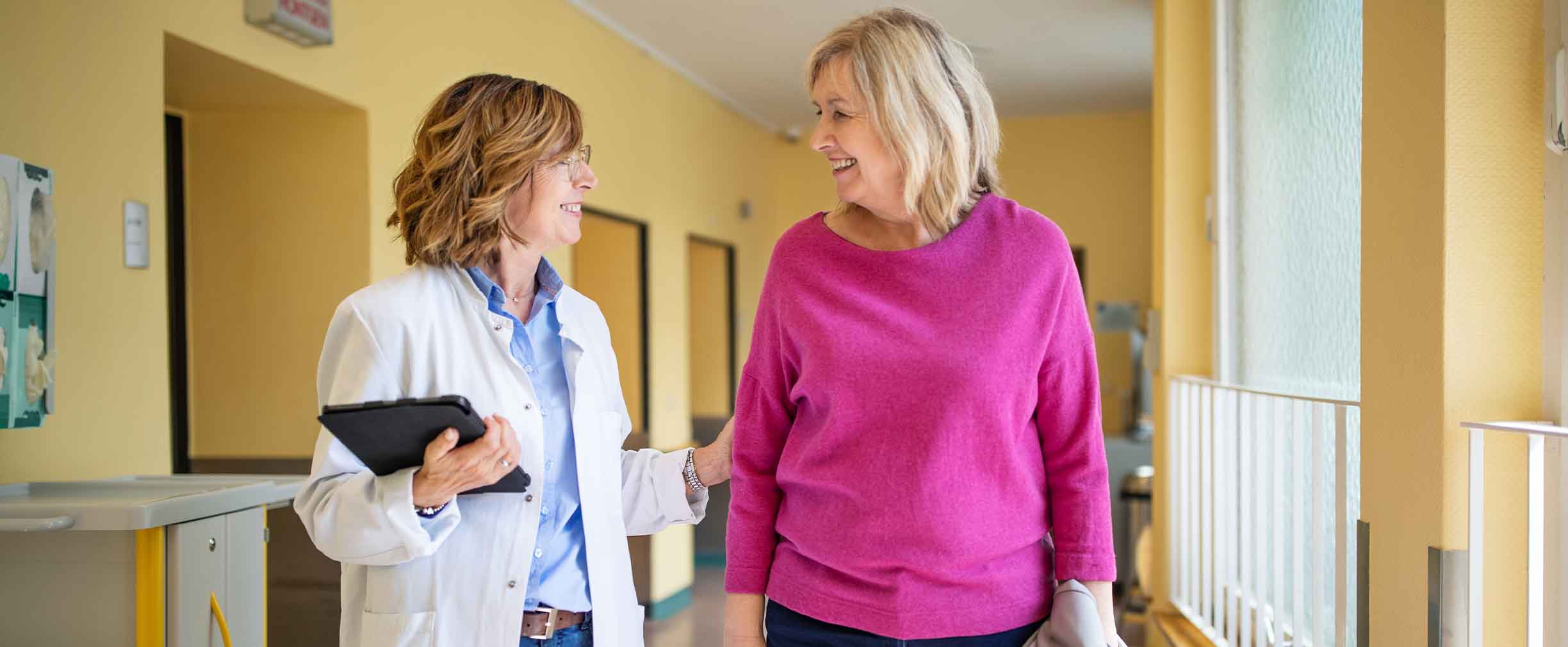 Una médica con bata de laboratorio blanca hablando con su paciente mientras camina por el pasillo.