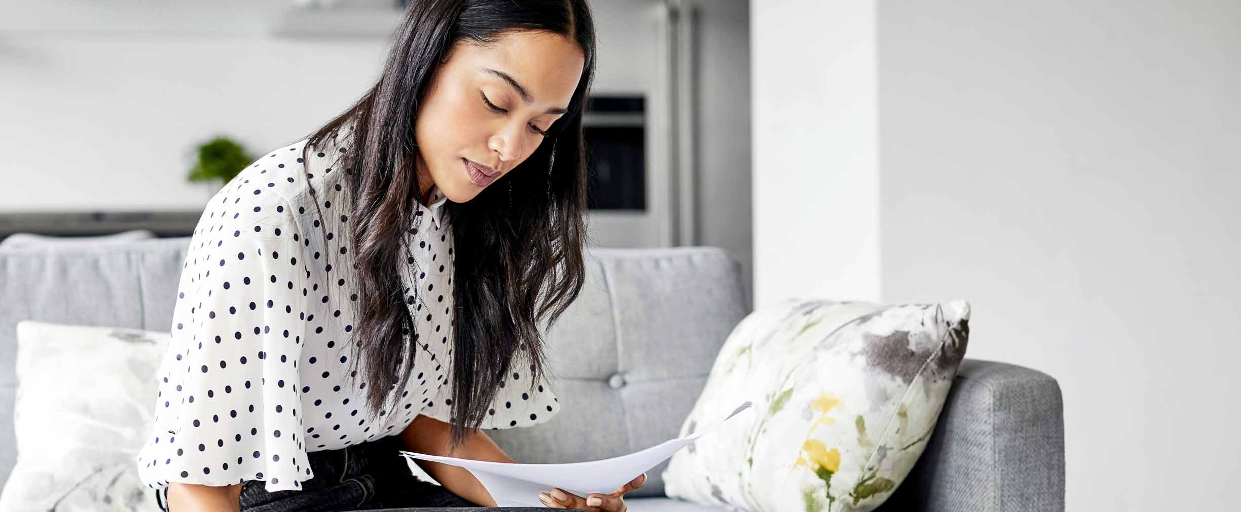 Mujer joven analiza facturas mientras escribe en una agenda. Mujer utiliza una tableta digital en la mesa. Está sentada sobre un sofá en casa.