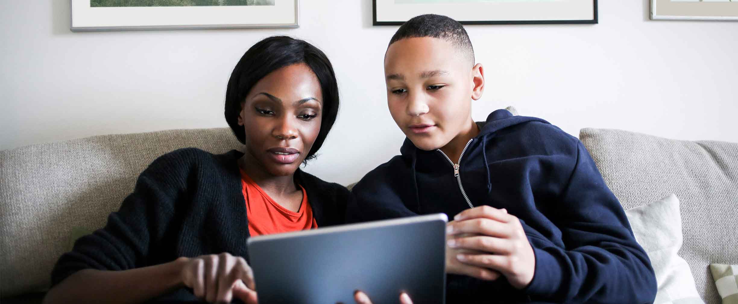 A mother sits on a sofa with her teenage child and they look at a tablet together.